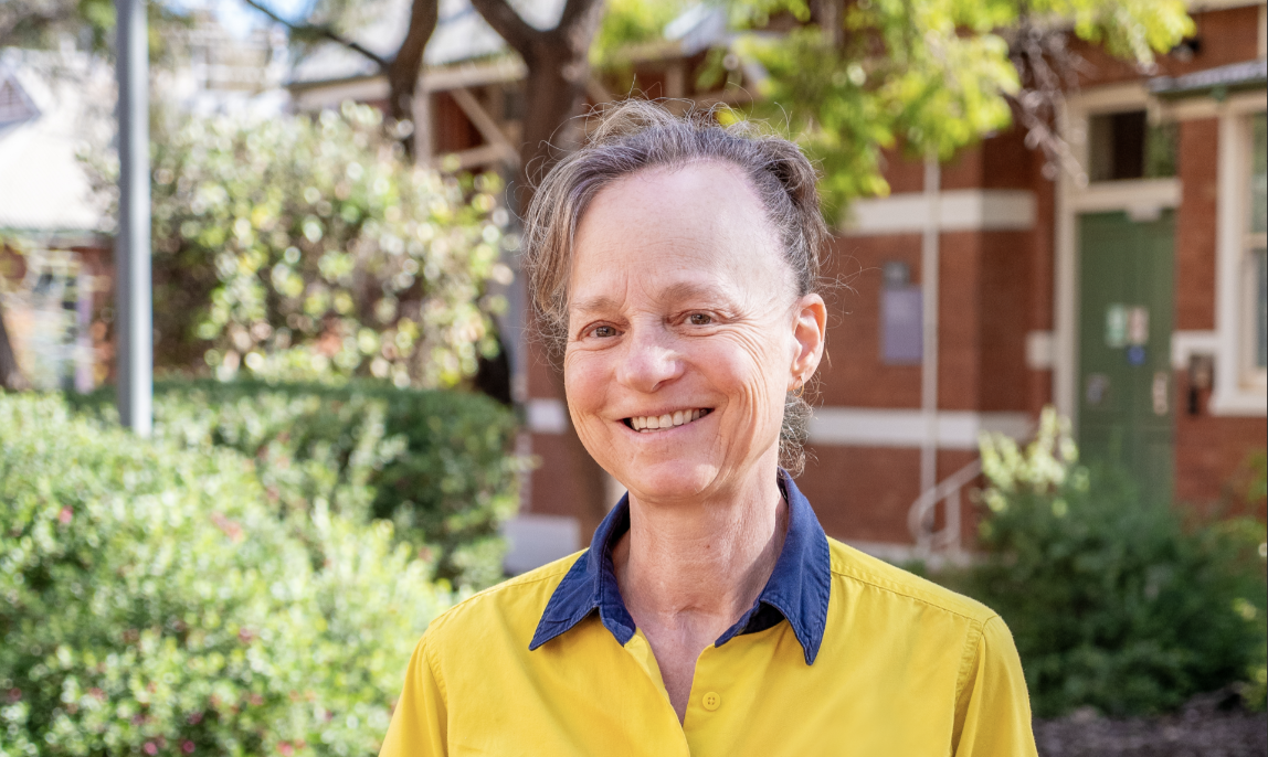 Student geologist Yeeda stand smiling on campus in front of a brick building and greenery. She wears her work uniform with her hair tied back.